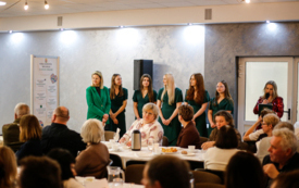 Publiczność siedząca na krzesłach i kobiety stojące w zielonych strojach i czerwonym garniturze.