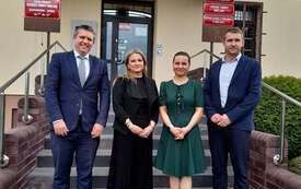 Przed budynkiem są cztery osoby. Dwie kobiety i dw&oacute;ch mężczyzn. Jedna z kobiet ubrana w czarną sukienką, adruga w zieloną. Mężczyźni w granatowych garniturach.