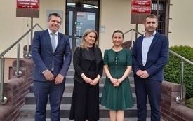 Przed budynkiem są cztery osoby. Dwie kobiety i dw&oacute;ch mężczyzn. Jedna z kobiet ubrana w czarną sukienką, adruga w zieloną. Mężczyźni w granatowych garniturach.