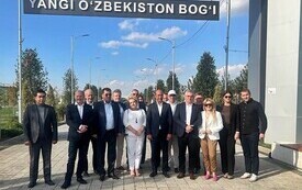 Na zdjęciu grupa przedstawicieli firm i samorząd&oacute;w stojąca na chodniku. U g&oacute;ry znak z napisem YANGI O'ZBEKISTON BOG'I.