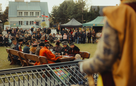Zdjęcie przedstawia mężczyzn w przebraniach siedzących przy drewnianym stole. W tle publiczność.