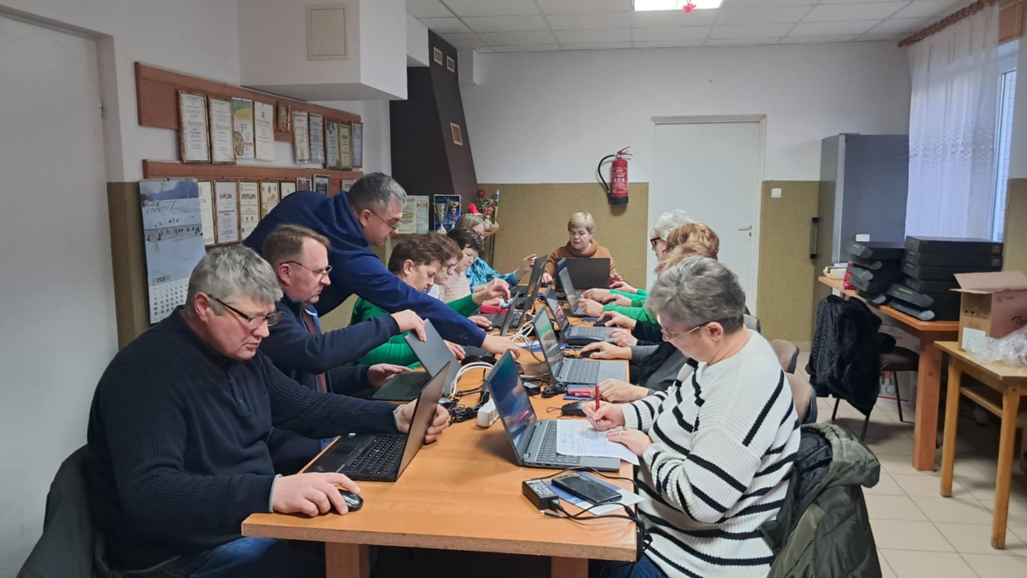 Kobiety i&nbsp;mężczyźni siedzący przy stolikach na&nbsp;kt&oacute;rych stoją laptopy. Jedna kobieta pisze coś na&nbsp;kartce.