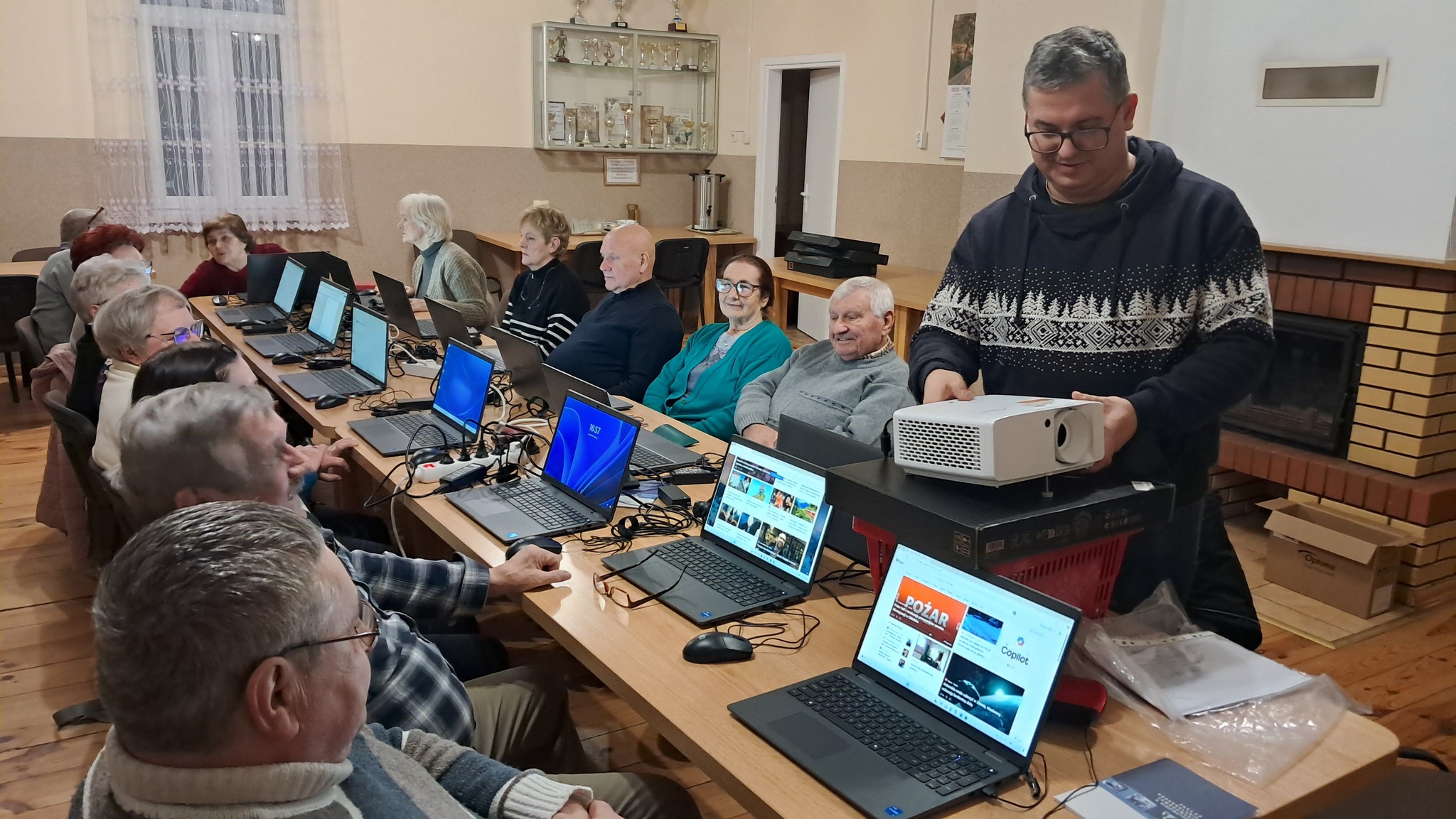 Kobiety i&nbsp;mężczyźni siedzący przy stolikach. Przed nimi stoją laptopy. Jeden mężczyzna ustawia sprzęt do wyświetlania na&nbsp;ścianie.