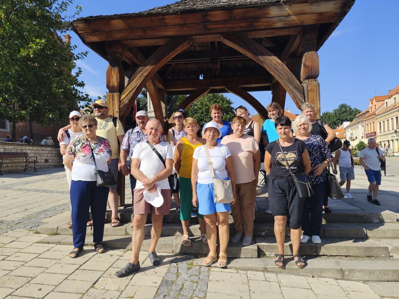 Osoby w letnich strojach stojące pod drewniana wiatką.