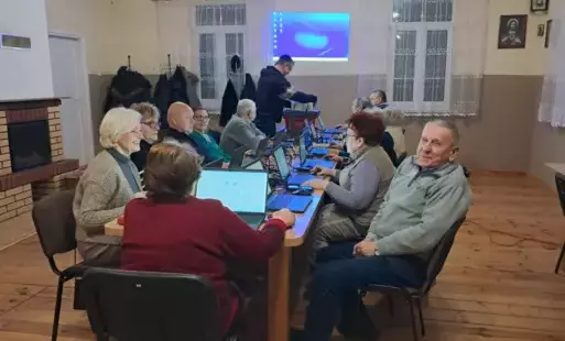 Kobiety i mężczyźni siedzący przy stolikach. Przed nimi stoją laptopy. Jeden mężczyzna ustawia sprzęt do wyświetlania na ścianie.