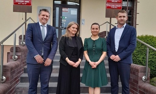 Przed budynkiem są cztery osoby. Dwie kobiety i dwóch mężczyzn. Jedna z kobiet ubrana w czarną sukienką, adruga w zieloną. Mężczyźni w granatowych garniturach.