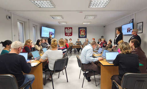Na zdjęciu, w sali, grupa urzędników podczas szkolenia. Przed każdym laptop. W tle na ścianie godło Polski oraz herb baranka na czerwono-zielonym tle.